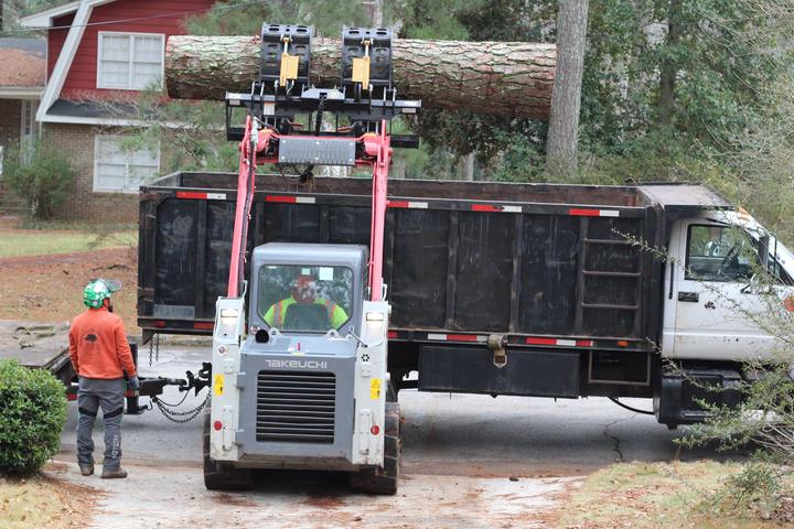Skid steer grapple lifting a long pine log over the dump truck while a crew member in an orange company shirt watches