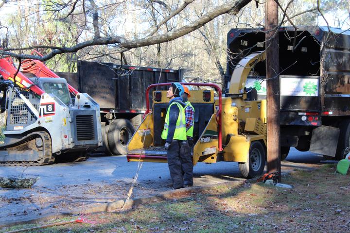 Two crew members in hard hats and safety vests looking up into the tree beside the chipper, with the skid steer and dump truck behind